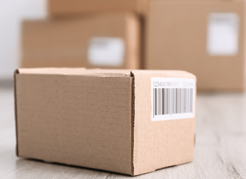A cardboard package with a prominent barcode on its side sits in the foreground, with a stack of similar packages blurred in the background, highlighting a focus on individual parcel tracking and delivery services offered by MyFreightWorld.
